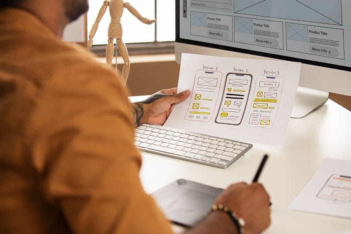 A designer at a desk reviewing three mobile wireframe sketches while referencing a desktop screen displaying website wireframes.
